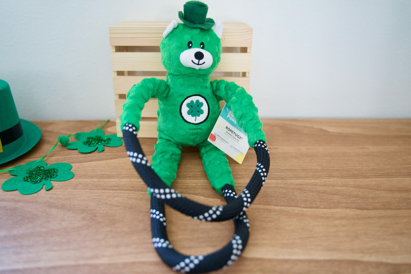 Un ours en peluche vert, coiffé d'un haut-de-forme et orné d'un trèfle, est posé sur une table en bois avec un jouet pour chien en corde.