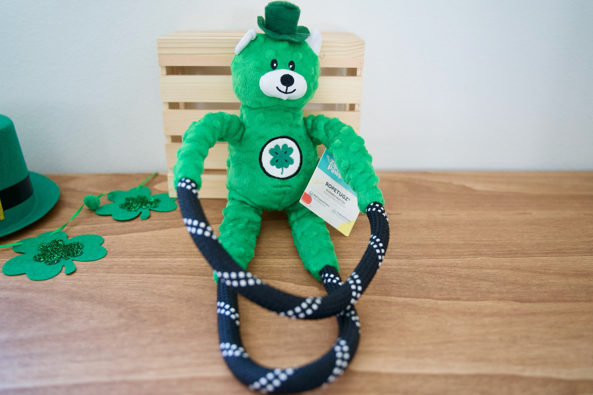 Un ours en peluche vert, coiffé d'un haut-de-forme et orné d'un trèfle, est posé sur une table en bois avec un jouet pour chien en corde.