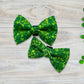 Two green dog bow ties with shamrock patterns on a wooden surface with clover leaves.