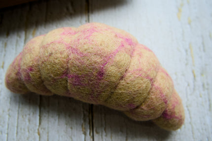 A handcrafted wool croissant dog toy with a beige body and pink swirls, resting on a white wooden surface.