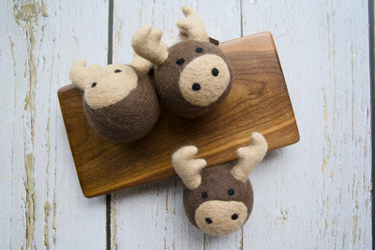 Three felted wool moose dog toys on a rustic wooden board.|Trois jouets pour chiens en forme d'élan, en laine feutrée, posés sur une planche en bois rustique.