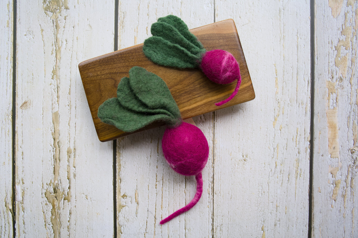 Two felted dog and cat toys shaped like radish with green tops and pink roots on a wooden board.