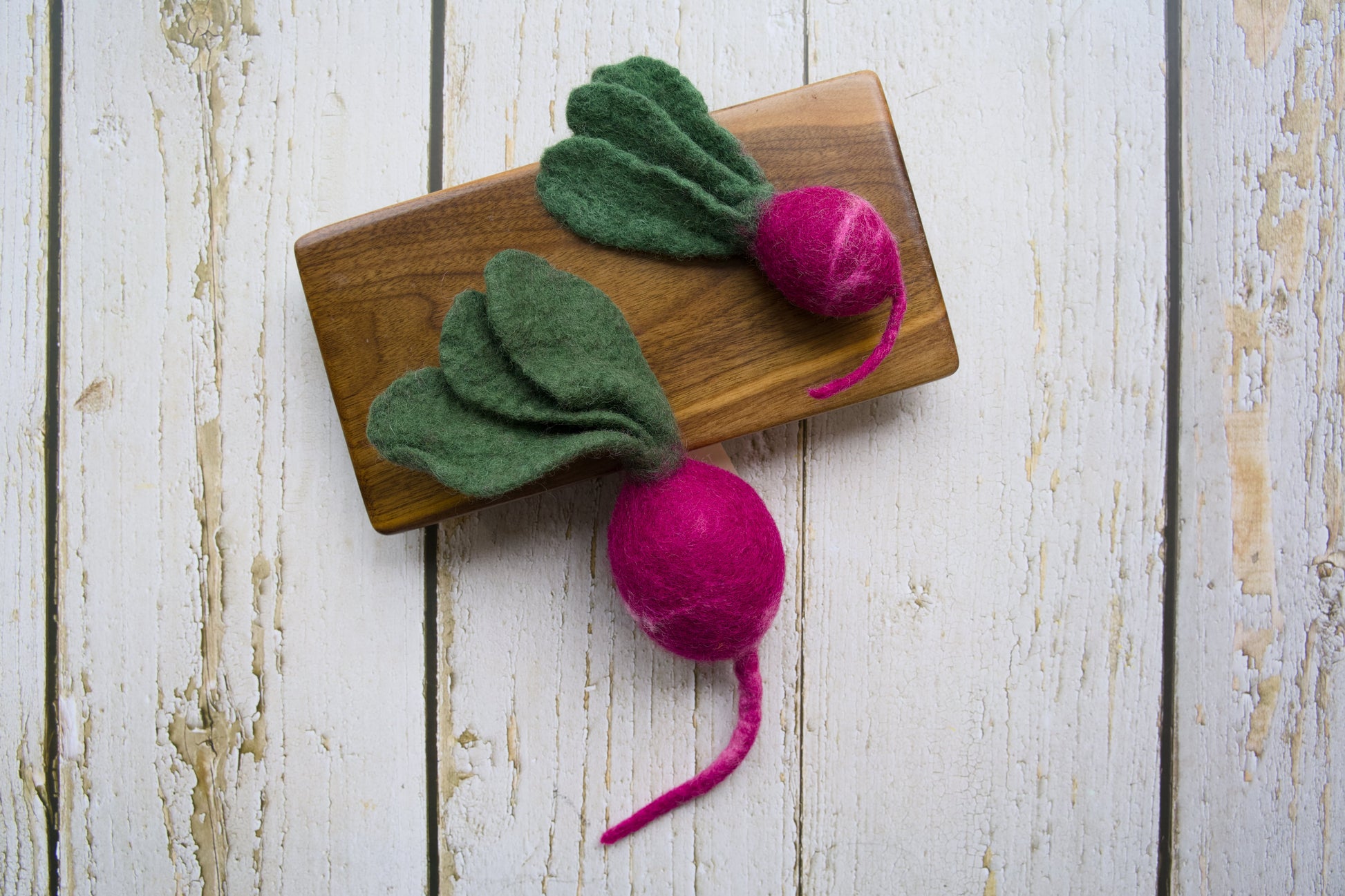 Two felted dog and cat toys shaped like radish with green tops and pink roots on a wooden board.