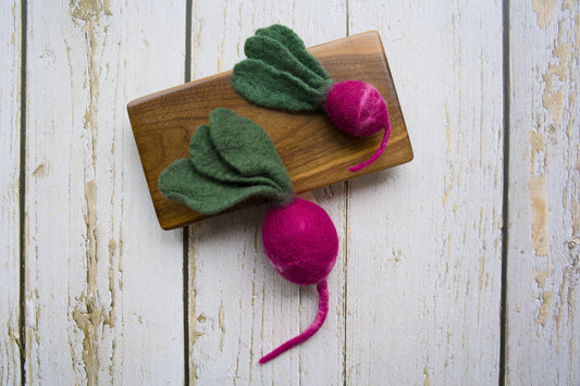 Two felted dog and cat toys shaped like radish with green tops and pink roots on a wooden board.|Deux jouets feutrés pour chiens et chats en forme de radis, avec des fanes vertes et une racine rose posé sur une planche en bois.