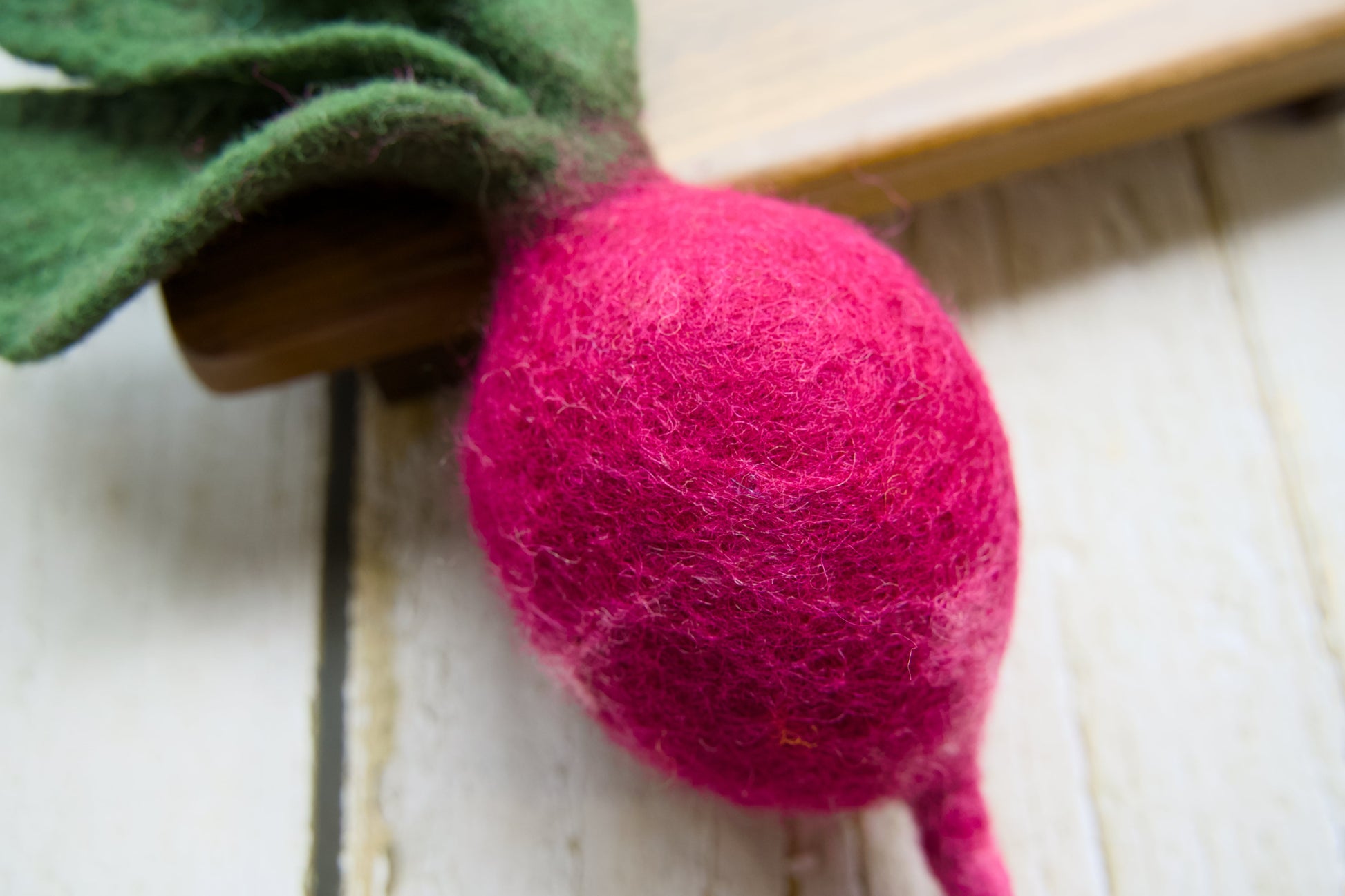 A close-up of a felted radish with a bright pink body and green felted leaves, resting on a white wooden surface.