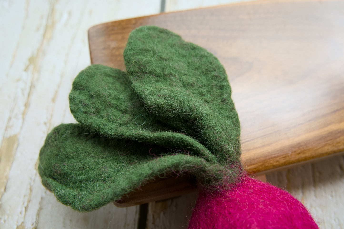 A close-up of a felted radish with green leaves and a bright pink root, resting on a wooden board.