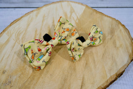 Deux nœuds papillon pour animaux à motifs de boissons colorées, posé sur une surface en bois.