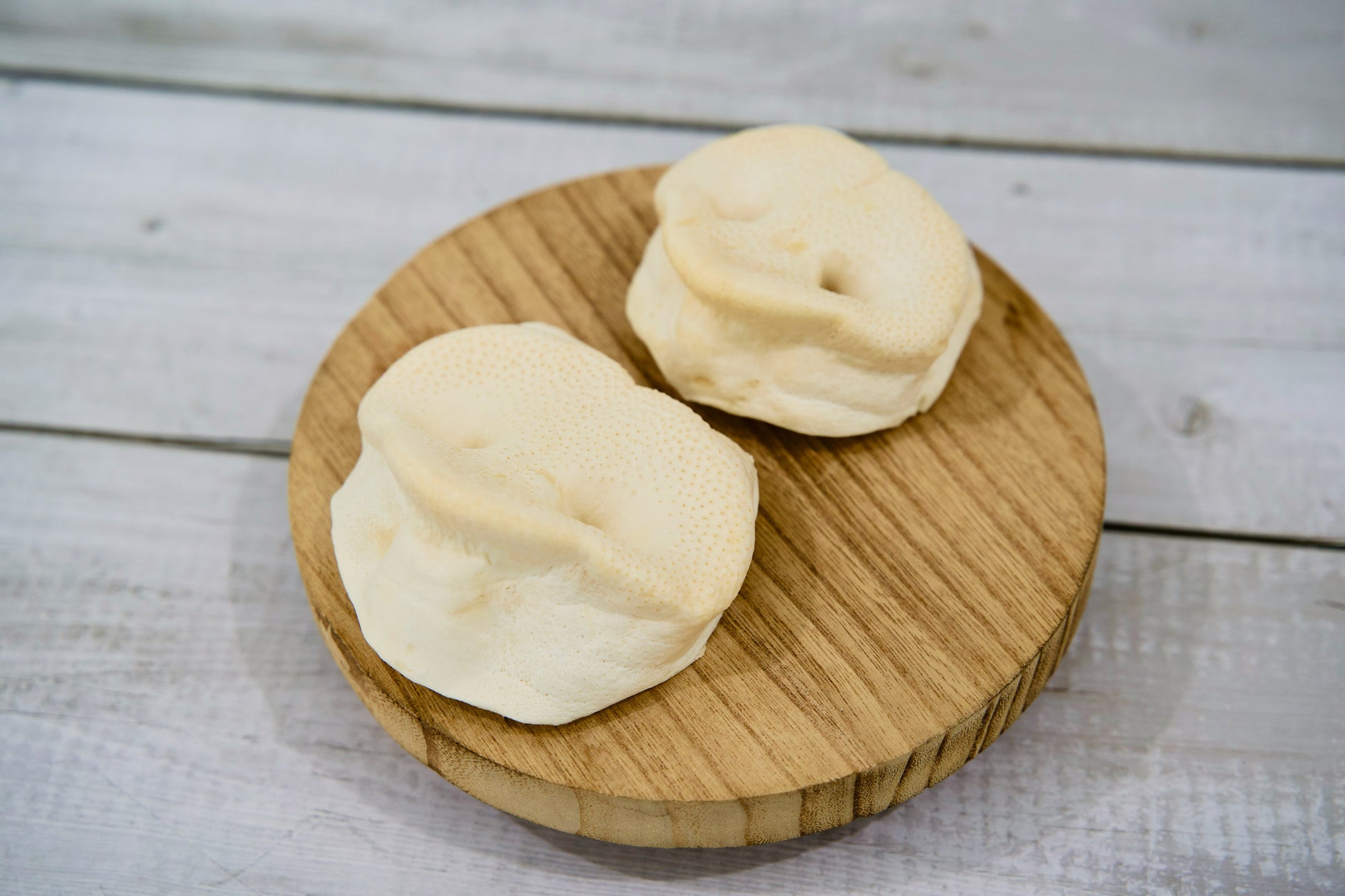 Deux friandises naturelles à mâcher pour chiens, à base de museaux de porc soufflés, présentées sur une planche ronde en bois, mettant en valeur leur texture croustillante et leur aspect entièrement naturel.
