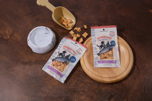 Two packages of Salmon Poke dog and cat treats are displayed on a dark brown textured surface.|Deux paquets de friandises « Salmon Poke » pour chiens et chats sont présentés sur une surface texturée brun foncé.