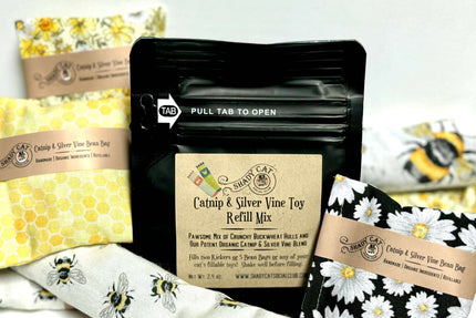 Un sachet noir refermable portant l'étiquette « Mélange de recharge pour jouets à l'herbe à chat et à la vigne argentée », entouré de coussins et de couchages douillets pour chats.