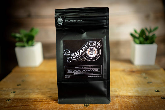 A black stand-up pouch of Shady Cat Social Club Fine Ground Organic Catnip tea, standing on a rustic wooden table.|Un sachet souple noir de tisane d'herbe à chat bio finement moulue « Shady Cat Social Club », posé sur une table en bois rustique.