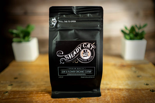 A black stand-up pouch of Shady Cat Social Club Tea & Flowers Organic Catnip stands on a rustic wooden table.|Un sachet souple noir de cataire biologique « Tea & Flowers » de Shady Cat Social Club est posé sur une table en bois rustique.