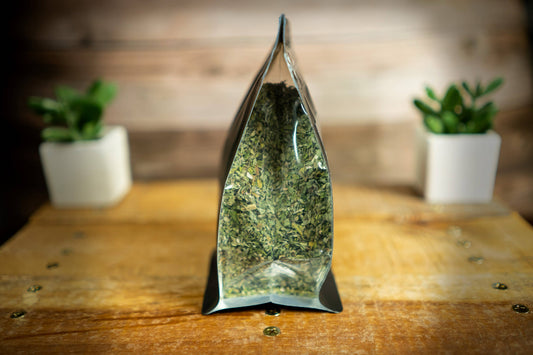 A clear stand-up pouch filled with dried leaf and flower catnip, standing on a wooden surface.|Un sachet souple transparent tenant debout, rempli de feuilles et de fleurs de cataire séchées, posé sur une surface en bois.