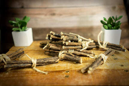 Des fagots de bâtonnets de vigne argentée attachés avec de la ficelle sont posés sur une surface en bois, avec des plantes en arrière-plan.
