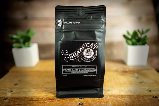 Organic catnip and silvervine blend bag labeled Shady Cat on a wooden surface with plants in the background.|Sachet de mélange d'herbe à chat biologique et de vigne argentée, étiqueté Shady Cat, posé sur une surface en bois avec des plantes en arrière-plan.