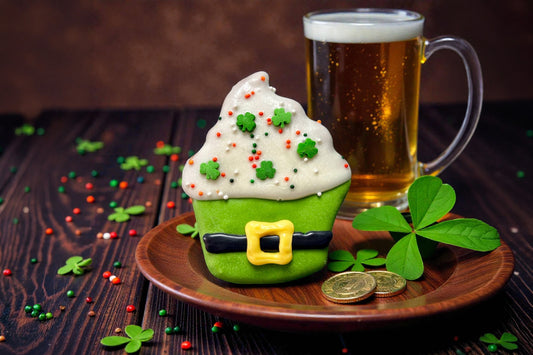 Green cupcake for dogs with white frosting, gold buckle, and shamrock decorations on a wooden plate.|Cupcake vert pour chiens avec glaçage blanc, boucle dorée et décorations en forme de trèfle, présenté sur une assiette en bois.