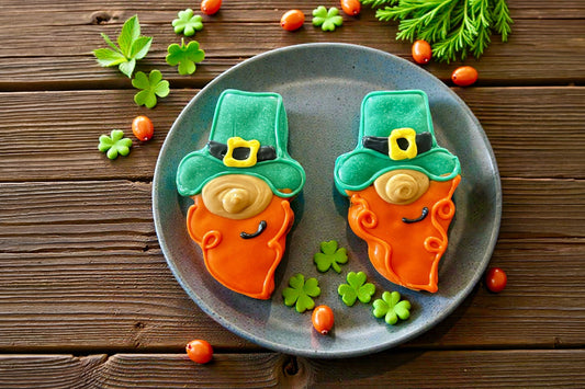 Two leprechaun-shaped cookies for dog on a plate with shamrock leaves.|Deux biscuits en forme de lutin pour chien, présentés sur une assiette décorée de feuilles de trèfle.